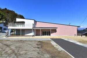 青空の下、砂利敷きにある、外階段とバルコニー付きの白とピンクの建物の斜め前からの写真。