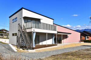 砂利と舗装路が手前にある、バルコニーと屋根付きエントランスを備えた白とピンクの建物の正面全景写真。