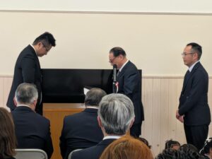 マイクの前で話す男性と見守る人々。